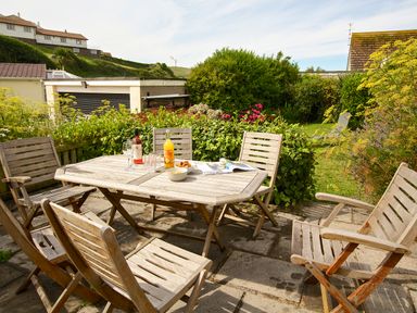 An outdoor patio with a wooden table and six chairs surrounded by bushes and flowers at Cindys Retreat in Hope Cove