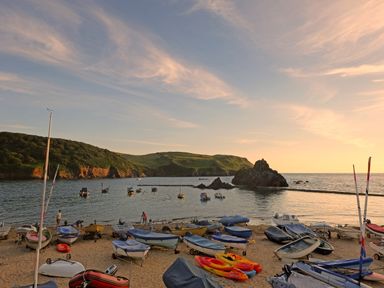 A beach with multiple small boats and kayaks on the sand by a calm sea with hills in the background at Cindy's Retreat in Hope Cove