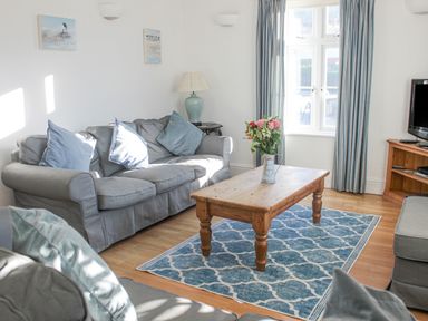 A living room with gray sofas a wooden coffee table flowers on the table a blue patterned rug and a window with curtains at Cindy's Retreat in Hope Cove