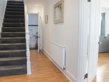 A hallway with wooden flooring a staircase with dark carpet and an open door leading to a sink and a room with a gray sofa at Cindy's Retreat in Hope Cove