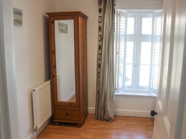 A small room with a wooden wardrobe with a mirror door a radiator a window with shutters and curtains and a door with a black handle at Cindys Retreat in Hope Cove