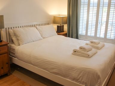 A bedroom with a white bed with pillows and folded towels on it next to two wooden bedside tables with lamps and white window shutters at Cindy's Retreat in Hope Cove