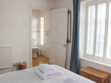 A bedroom with folded towels on a bed an open door showing a bathroom with a toilet a window with shutters and a wicker basket at Cindy's Retreat in Hope Cove