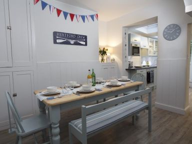 A dining area with a wooden table set with plates and glasses and blue chairs next to a kitchen at Crab Cottage Salcombe
