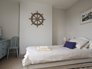 A bedroom with a bed with white bedding and a blue cushion a wooden chair a chest of drawers a nautical wheel on the wall and a framed picture at Crab Cottage in Salcombe