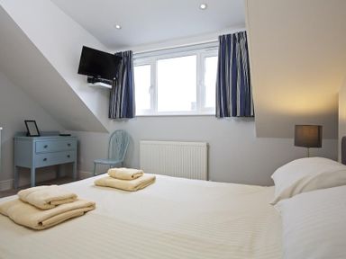 A bedroom with a bed towels on the bed a bedside lamp a blue chair a blue chest of drawers a TV mounted on the wall and windows with striped curtains at Crab Cottage in Salcombe