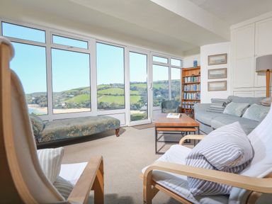 A sitting room with chairs a sofa a coffee table and a bookcase with large windows overlooking green hills at Edinburgh House in Salcombe