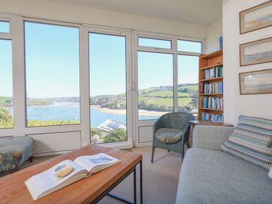 A living room with a bookshelf sofa chair and coffee table with an open book by large windows overlooking a river and hills at Edinburgh House in Salcombe