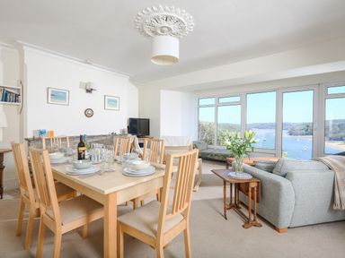 A dining area with a wooden table set for six and a sitting area with a sofa and large windows overlooking a river at Edinburgh House in Salcombe