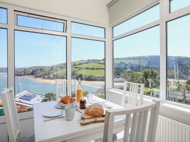 A dining area with a white table and chairs set with bread croissants orange juice and mugs overlooking a river and hills at Edinburgh House in Salcombe