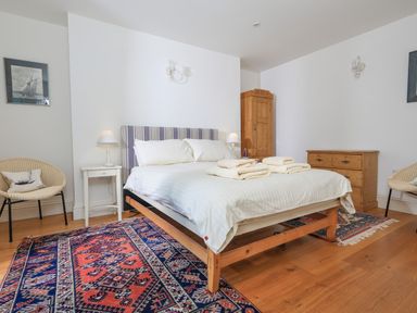 A bedroom with a bed on a wooden frame bedside tables lamps a wooden wardrobe and chest of drawers with chairs on a rug at Edinburgh House in Salcombe