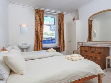 A bedroom with two beds white linens a wooden dresser and a window with patterned curtains at Edinburgh House in Salcombe