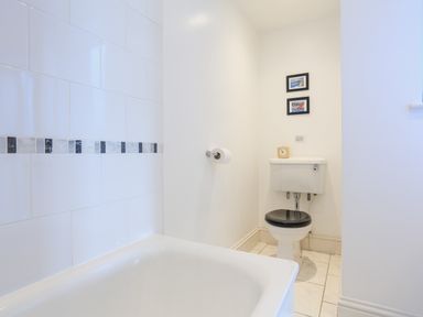 A bathroom with a bathtub tiled wall and a toilet with a black seat and framed pictures above at Edinburgh House in Salcombe