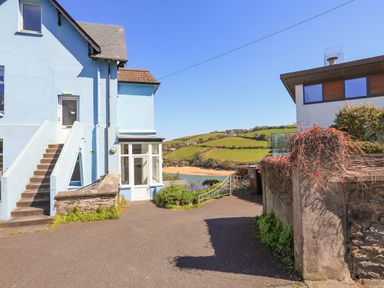 Exterior view of a blue painted house with stairs and a driveway overlooking green hills and a beach at Edinburgh House in Salcombe
