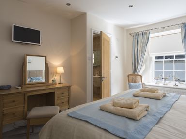 A bedroom with a bed folded towels on a blue runner a wooden vanity desk with mirror and lamp a chair by the window with curtains and ornamental birds on the sill at Gooder House in Dartmouth