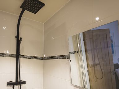 A bathroom corner with a black shower fixture and white tiled walls with a mirror reflecting a wooden door at Gooder House in Dartmouth