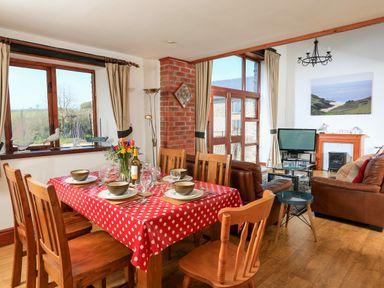 A dining area with wooden chairs and a table with red polka dot tablecloth next to a living room with leather sofas a TV and a fireplace at The Granary Malborough