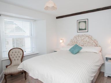 A bedroom with a bed a patterned headboard a chair by a window with blinds and bedside tables at Hazel Cottage in Malborough