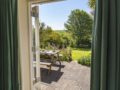 A garden with a wooden picnic table on a stone patio viewed through open doors with green curtains at Hazel Cottage Malborough