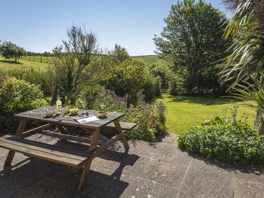 A wooden picnic table with bowls and drinks on a stone patio overlooking a garden with trees and a grassy field at Hazel Cottage in Malborough