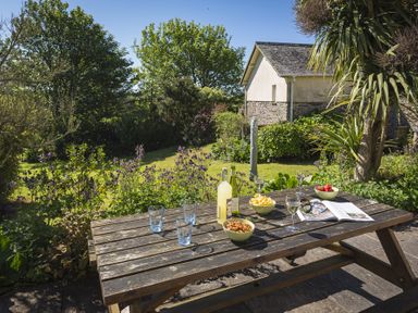 An outdoor wooden picnic table with drinks snacks and an open magazine in a garden at Hazel Cottage in Malborough