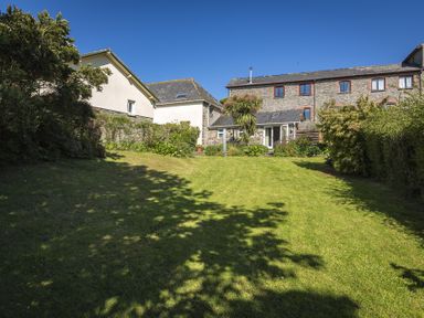 A backyard with grass lawn and shrubs behind a stone and white-walled house at Hazel Cottage in Malborough