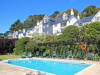 An outdoor swimming pool with lounge chairs and a pergola in front of residential buildings surrounded by trees and shrubs at The Hoot 16 St Elmo Court in Salcombe