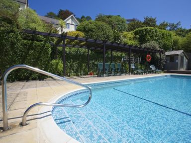 An outdoor swimming pool with a metal handrail next to a pergola with green chairs and a small building in the background at The Hoot 16 St Elmo Court Salcombe
