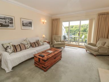 A living room with a sofa, armchairs, a wooden chest used as a table, wall art, and a sliding glass door leading to a balcony at The Hoot, 16 St Elmo Court in Salcombe