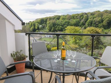 A balcony with a round metal table and four chairs with a bottle and four glasses on the table overlooking a wooded hillside at The Hoot 16 St Elmo Court in Salcombe