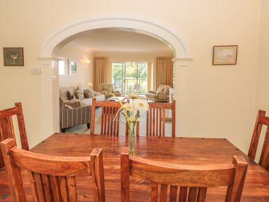 A dining room with wooden table and chairs and a vase of flowers looking through an arch into a living room with sofas and armchairs at The Hoot 16 St Elmo Court in Salcombe