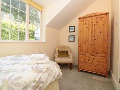 A bedroom with a bed with folded towels near a window an upholstered chair and a wooden wardrobe at The Hoot 16 St Elmo Court Salcombe