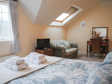 A bedroom with a bed, folded towels, a television, a sofa, a wooden desk with a mirror, and a window at The Hoot 16 St Elmo Court in Salcombe