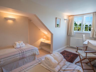 A bedroom with two single beds with folded towels desk chair window and small table lamp at Jot Cottage in Salcombe