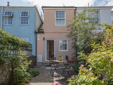 A small patio with a table and four chairs outside a two-story peach colored house with plants around at Jot Cottage in Salcombe