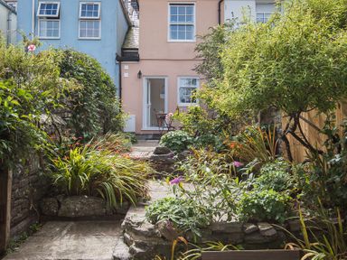 A garden path with plants and flowers leading to a pink house with a table and chairs at Jot Cottage in Salcombe