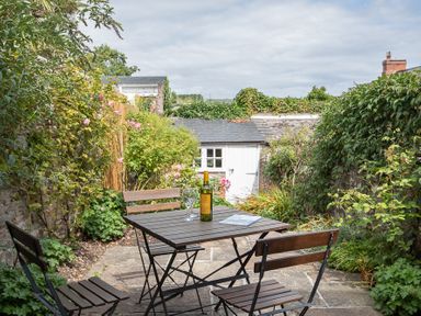 A small outdoor patio with a wooden table and chairs with a bottle and glasses surrounded by plants at Jot Cottage in Salcombe