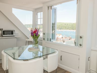 A dining area with a glass table and white chairs next to a large window overlooking a harbor at Kings View in Dartmouth