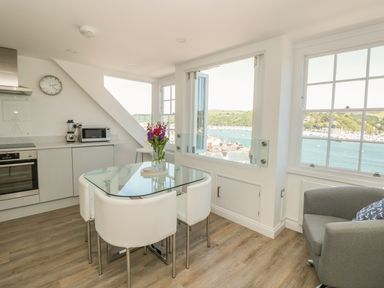 A kitchen dining area with glass table white chairs grey armchair and windows overlooking water at Kings View in Dartmouth