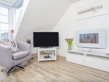 A living room with a grey armchair and pillow a television on a white stand a white cabinet with a flower vase and a wall-mounted fireplace at Kings View in Dartmouth