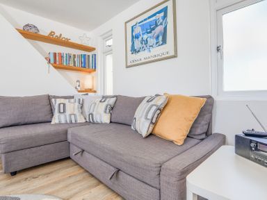 A living room corner with a gray sectional sofa with patterned and solid pillows a white side table with a radio wooden shelves with books and decor items and a framed artwork on the wall at Kings View in Dartmouth
