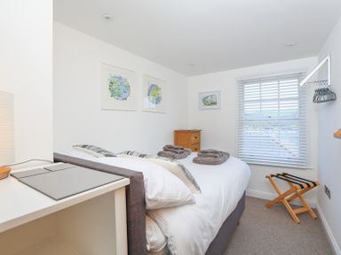 A bedroom with a double bed white bedding and folded towels a small wooden cabinet window with blinds and a luggage rack at Kings View in Dartmouth