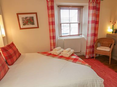 A bedroom with a bed with red plaid pillows and blanket facing a window with blinds and red check curtains at Lee Shore in Salcombe