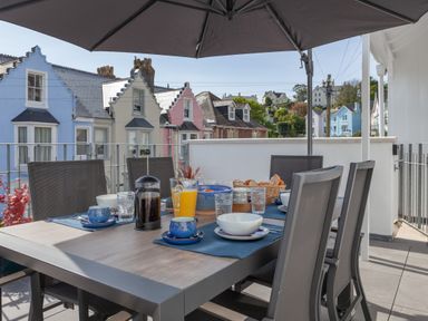 An outdoor dining table with chairs set for breakfast under a large umbrella on a balcony with colorful houses in the background at Lower Fernlea in Salcombe