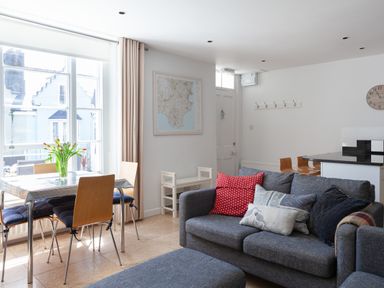 A living room with a gray sofa with cushions a dining table with chairs and flowers on it near a large window at Lower Fernlea in Salcombe