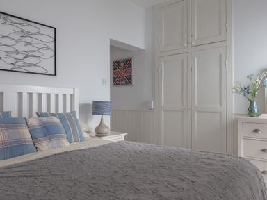 A bedroom with a white bed frame and blue plaid pillows a bedside table with a lamp built in cupboards and a dresser with flowers in a vase at Lower Fernlea in Salcombe