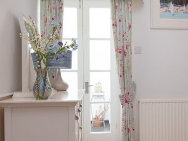 A dresser with flowers in a vase and a lamp near glass doors with floral curtains at Lower Fernlea in Salcombe