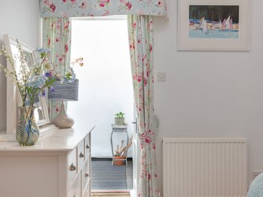 A room with a white dresser holding a vase with flowers and a lamp next to floral curtains and a small outdoor stand with plants at Lower Fernlea in Salcombe