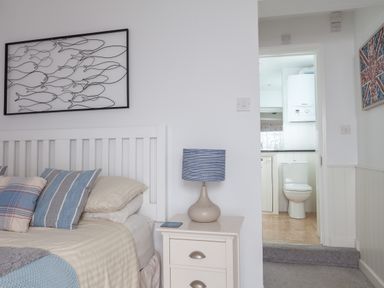 A bedroom with a white bed with striped pillows a bedside table with a lamp and a view into a bathroom at Lower Fernlea in Salcombe