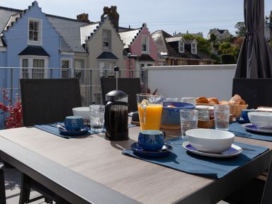 An outdoor dining table set with blue cups, bowls, glasses, a French press, orange juice, and a basket of bread on a terrace with houses in the background at Lower Fernlea in Salcombe